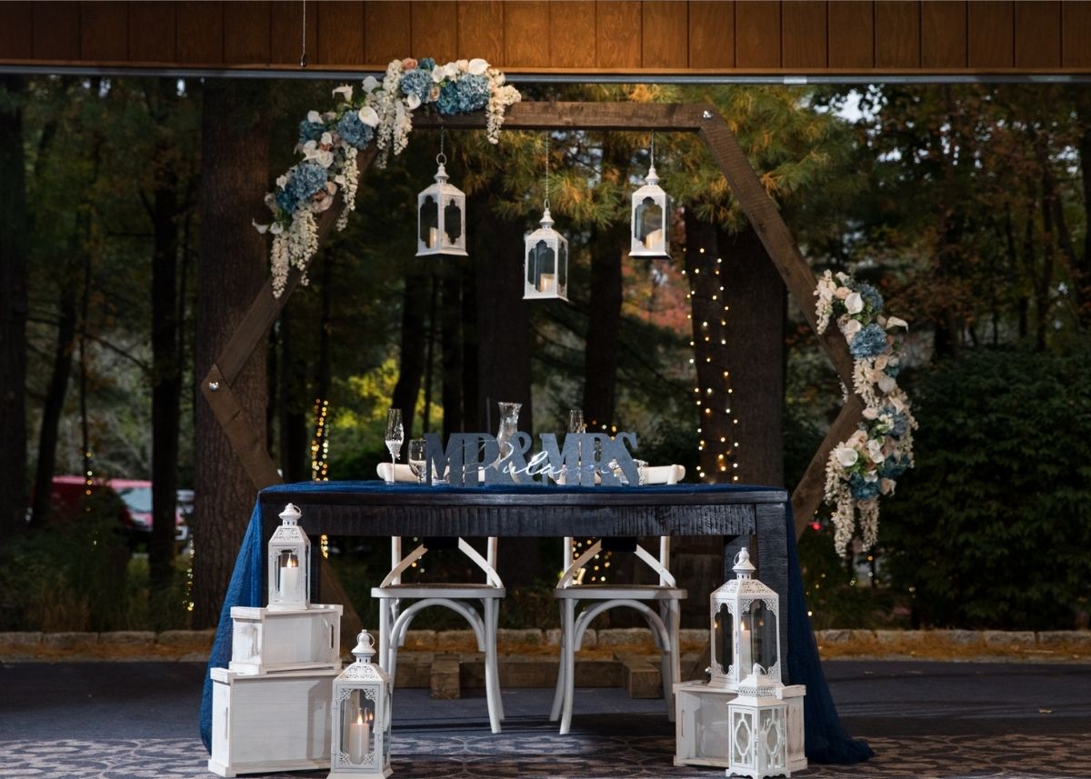 Custom personalized hexagon wedding arch with dark stained wood, metal hardware, and flowers on both corners, placed behind the bride and groom's table to create a stunning photo backdrop with lanterns and romantic decor
